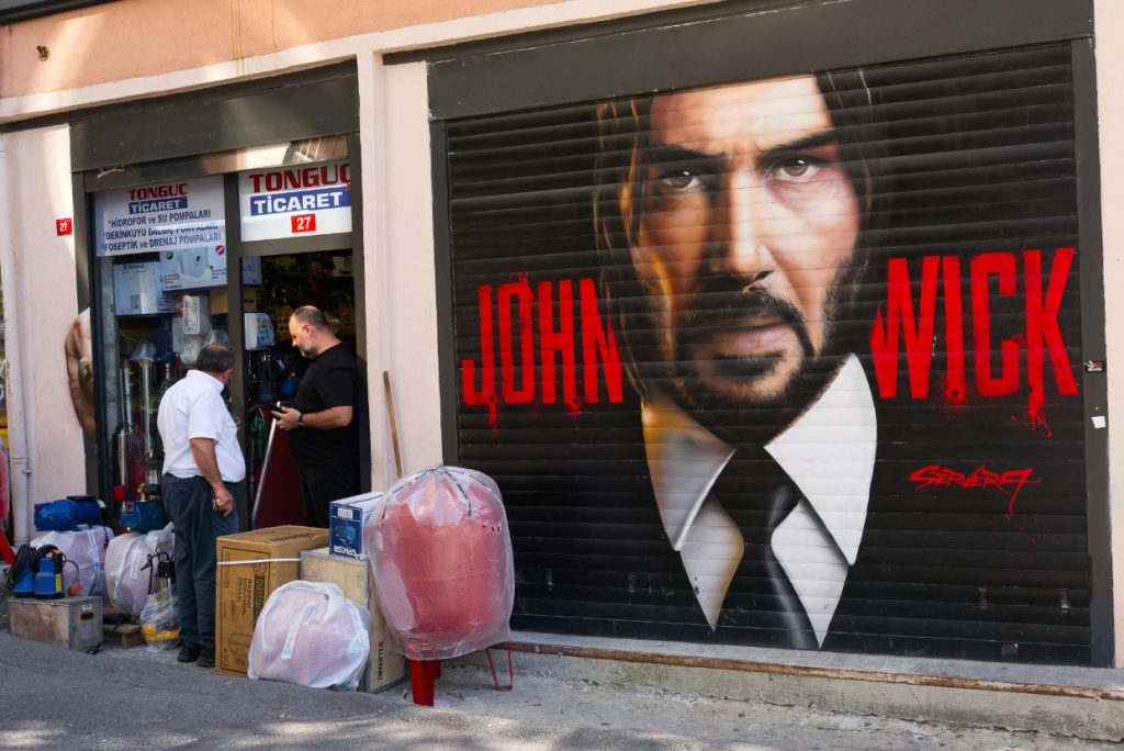 A large John Wick mural painted on a shop shutter in Istanbul, beside a local store displaying household goods on the pavement.
