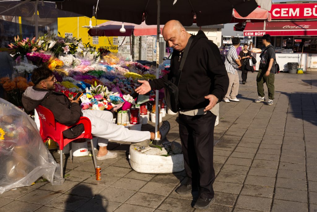 A man gestures while talking to a flower seller at a colourful outdoor flower stall on a sunny Istanbul street.