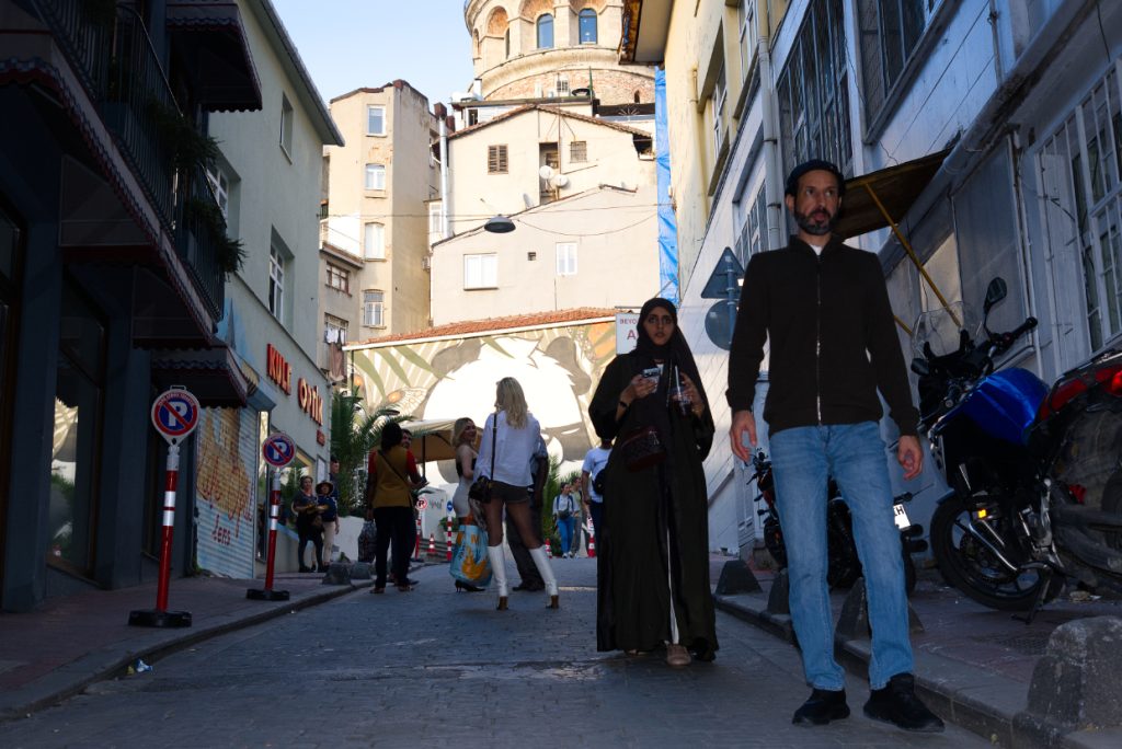 A man and woman walk along a narrow, shaded street in Istanbul, with Galata Tower rising above the sunlit buildings in the background.