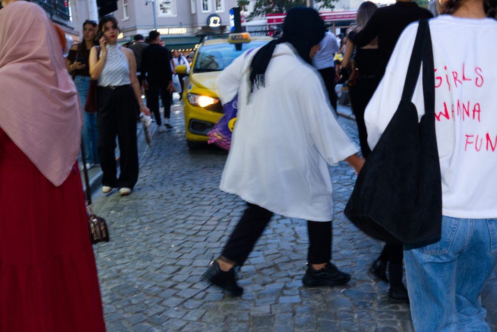 Pedestrians crossing a crowded cobblestone street near Galata Tower in Istanbul, with a yellow taxi approaching between shoppers and colourful storefronts.